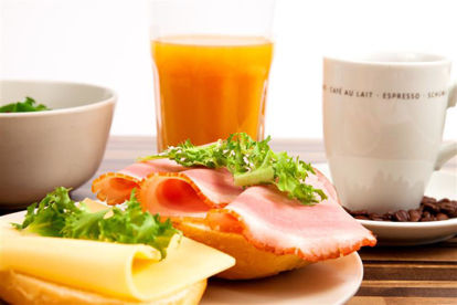 Das Bild zeigt ein belegtes Brötchen, im Hintergrund eine Tasse Kaffee und ein Glas Orangensaft